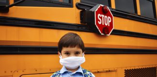 Coronavirus school reopening concept. A boy student stands in front of school bus wearing a face mask and backpack with stop sign clearly visible.