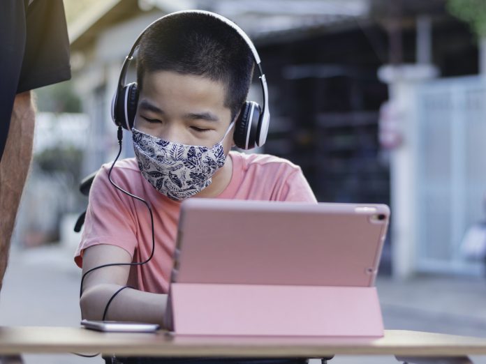 Happy asian disabled on wheelchair wearing Handmade mask to protection flu Covid 19 or Coronavirus, Stay at home to prevent the spread of the virus disease 2020, Boy uses his tablet to study online.
