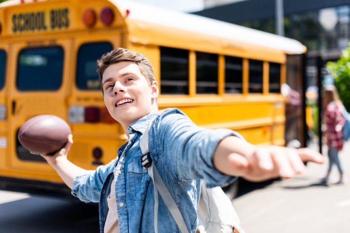 happy teen schoolboy throwing american football ball in front of school bus