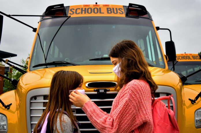 Female child helping another with a facemask in front of school bus