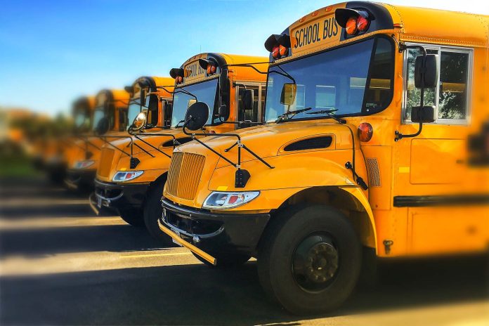 Row of yellow school buses parked inline with blurred background and sky reflecting off of windshields
