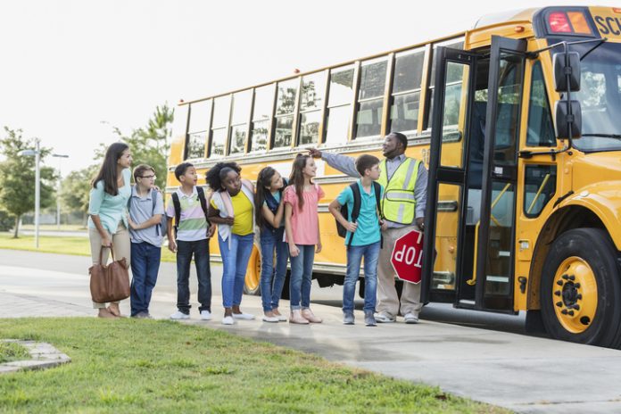 A multi-ethnic group of six middle school students, 11 to 13 years old, with their Hispanic teacher and a school crossing guard or bus driver, standing together outside a school bus, getting ready to go on a field trip. The boy wearing eyeglasses next to the teacher has down syndrome.