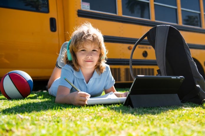 Pupil writing homework on grass with laptop tablet outdoor near school bus. Learning online, child education concept