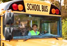 Portrait Of Happy Black Female Driver Driving Yellow School Bus, Beautiful Young African American Lady In Reflecting Vest Looking Through Window, Holding Steering Wheel An Smiling At Camera